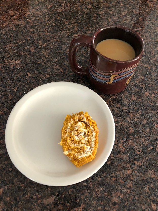 image of a slice of pumpkin spiced roll on a white plate, next to a mug of coffee